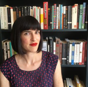 A woman is standing in front of a bookshelf filled with books. She is short black hair and wears red lipstick.