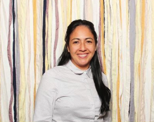 Maria Celleri, a Latinx woman with long dark hair, is smiling and wearing a light-colored button up shirt. She is standing in front of a wall with different colored veritcal stripes.