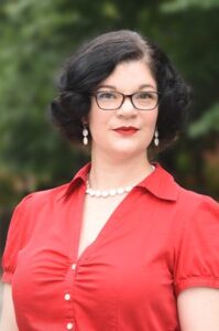 A white woman with short dark hair is standing outside in front of trees. She is wearing a red top and red lipstick. She is wearing a white-beaded necklace with a matching pair of earrings.