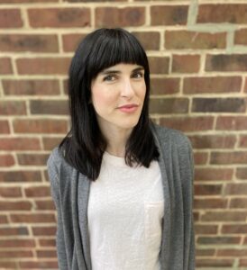 Keegan Cook Finberg, a white woman with shoulder-length dark hair and bangs, stands in front of a brick wall. She is wearing a white t-shirt with a gray cardigan on top.