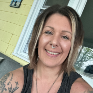 Jessica Floyd, a white woman with short brown hair and blonde highlights, is sitting on a porch and smiling at the camera. She has two nose piercings.
