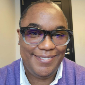 Tanya Saunders, a Black woman with short hair, is smiling at the camera. She is wearing eyeglasses and has on a purple sweater.