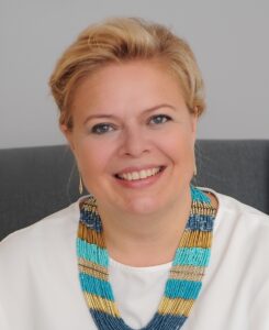 Irina Golubeva, a white woman with her blonde hair in an updo, is sitting in a gray chair and smiling at the camera. She is wearing a white blouse and a colorful necklace