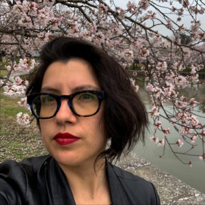 Tania Lizarazo, a Latinx woman with short brown hair, is standing in front of a cherry blossom tree. She has on eyeglasses and is wearing red lipstick.
