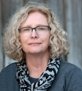 Carole McCann, a white woman with curly blond hair, is wearing eyeglasses. She looks directly at the camera.