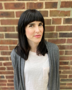 Keegan Cook Finberg, a white woman with shoulder-length dark hair and bangs, stands in front of a brick wall. She is wearing a white t-shirt with a gray cardigan on top.
